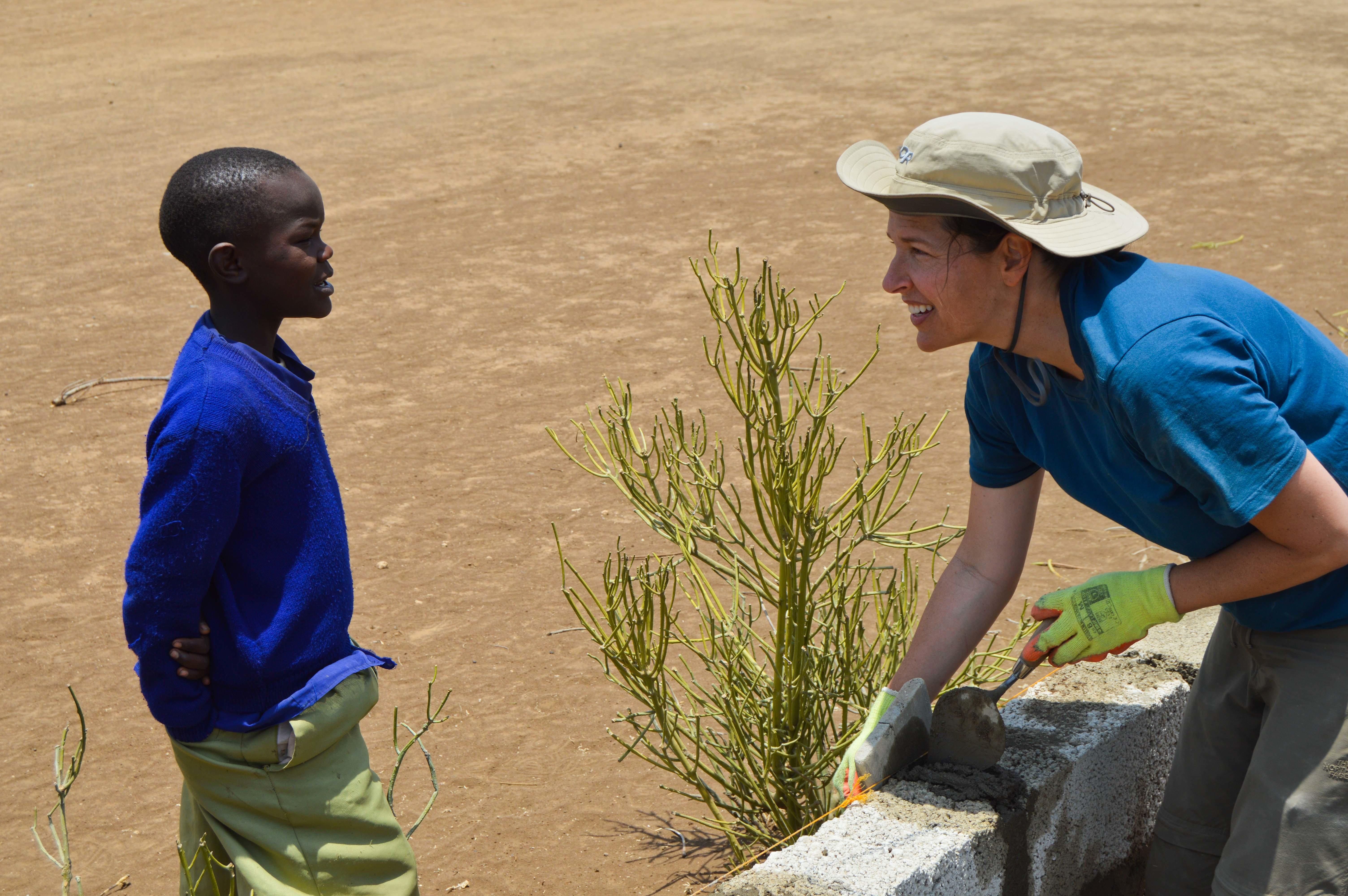 A Projects Abroad teenage building volunteer working in Tanzania chats with a young child while laying bricks. A teenage volunteer doing construction work in Tanzania builds a wall while chatting to a young child.